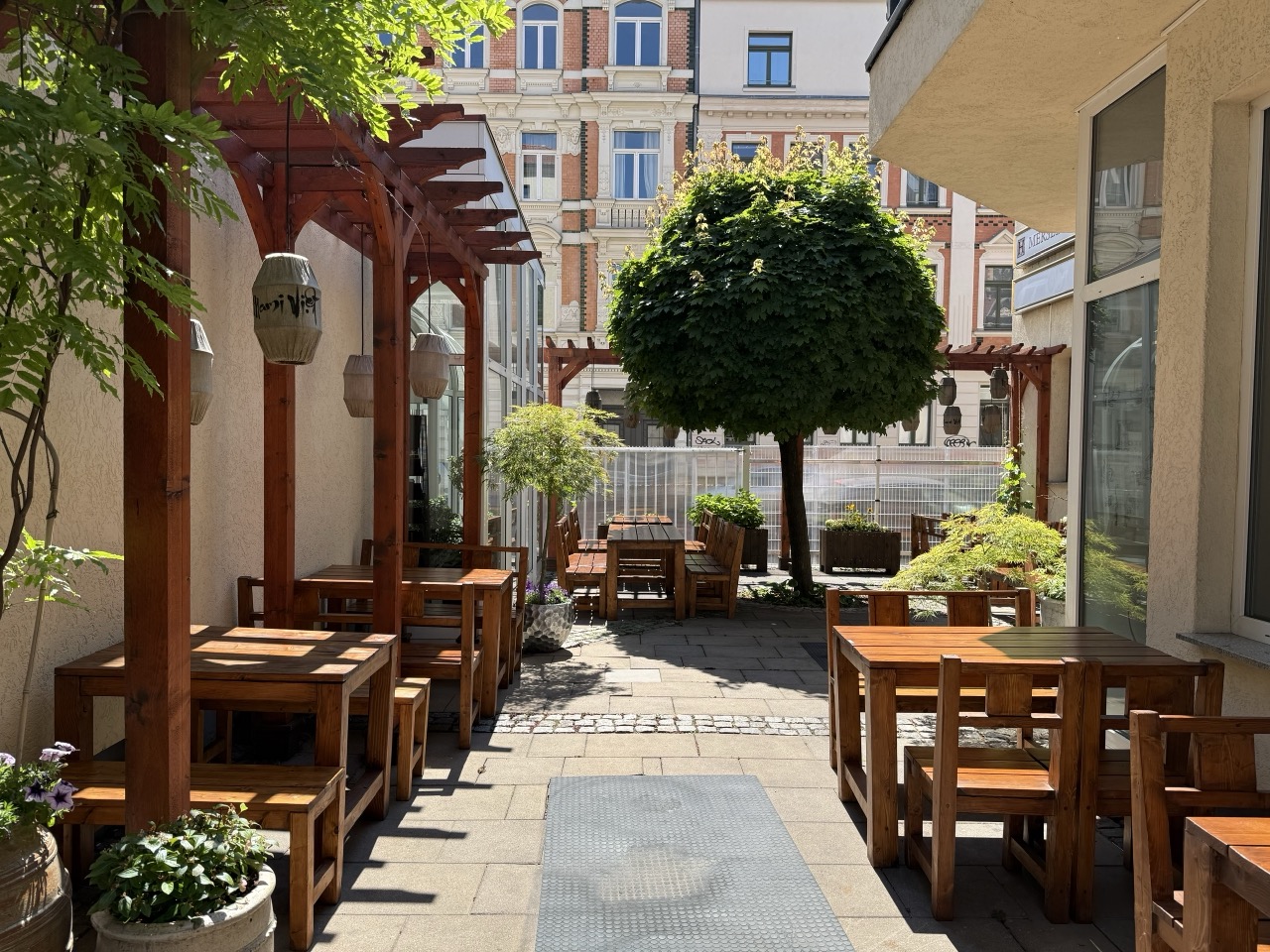 Sonniger Gartenbereich von Mami Viet in Leipzig-Lindenau mit Holztischen, Pergola und Lampions Sonniger Gartenbereich von Mami Viet in Leipzig-Lindenau mit Holztischen, Pergola und Lampions